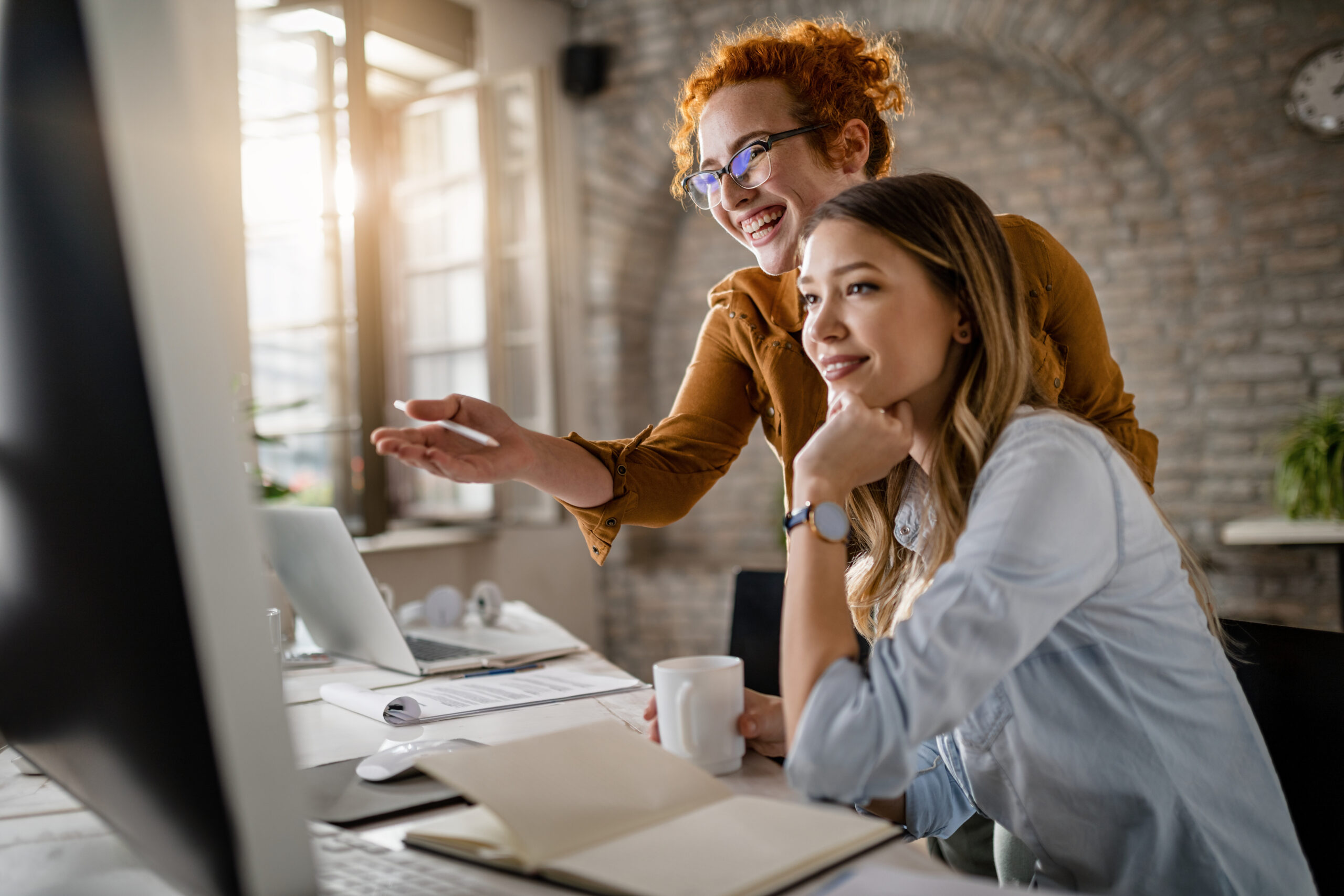 Happy female entrepreneurs reading an e-mail on computer while working together in the office. Focus is on redhead woman.